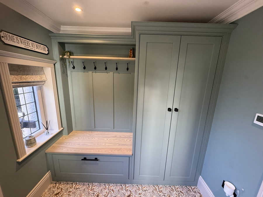 A sage green boot room with an oak bench top, hook rail and tall cupboards