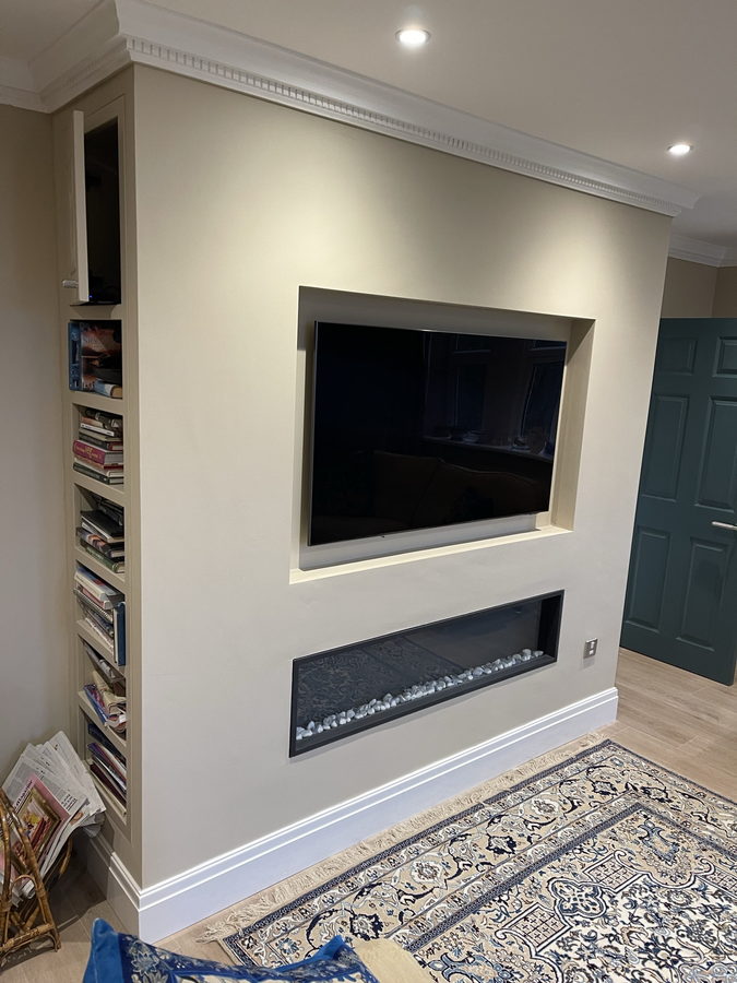 A cream panelled wall with a linear fire, book shelving and teal cupboards